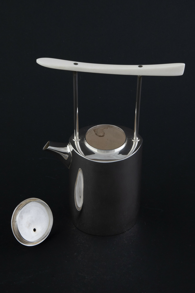 Kaffekanne i sølv. Ovalformet korpus med lett bølgende vegger og liten tut. Fra skulderen reiser det seg to tynne rør som forbinder kannen med et håndtak i elfenben. Sirkulært lokk med sylinderformet lokknapp i elfenben. 