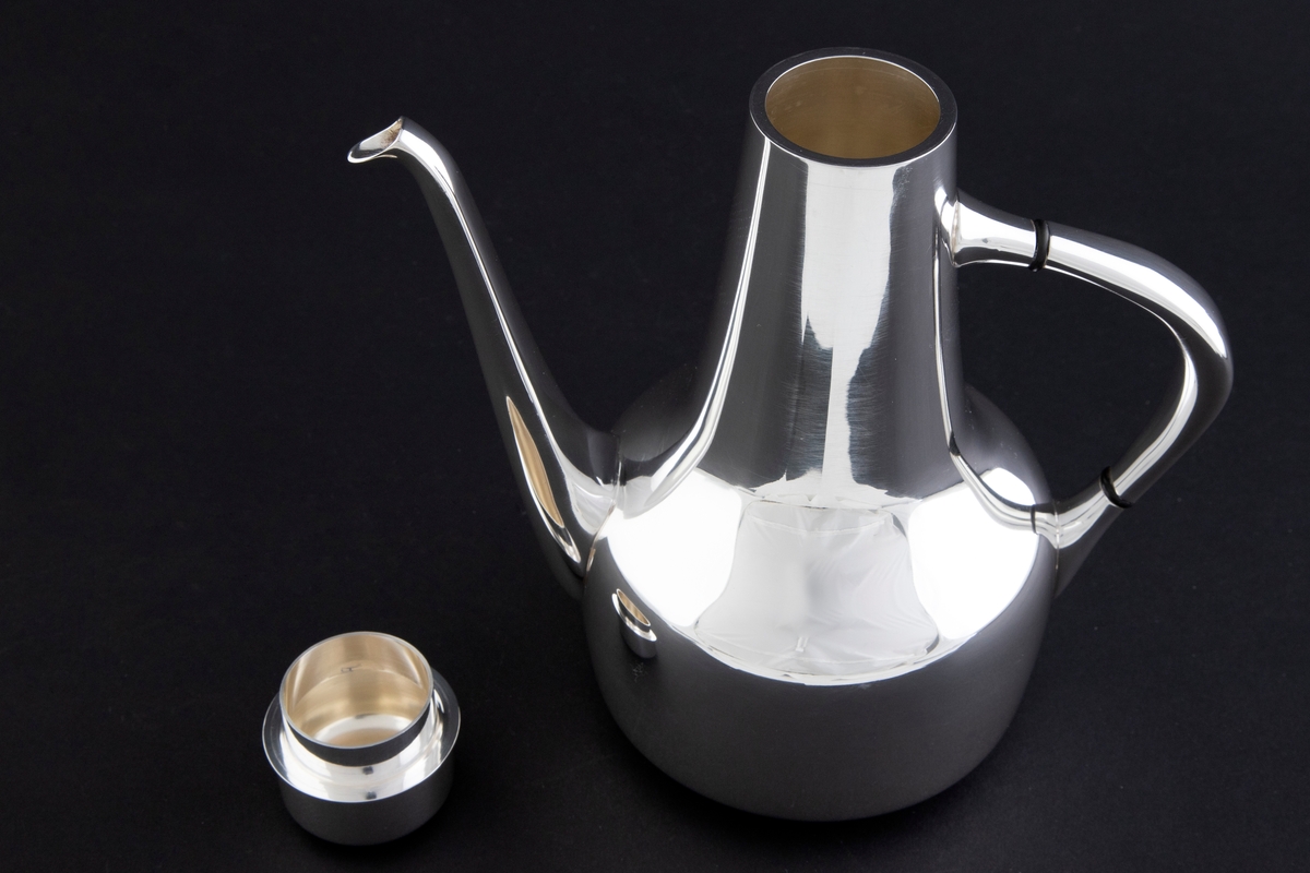 Kaffekanne som er utført i glattpolert sølv, uten dekor og med myke kanter. Sylinderformet kompakt korpus, som glir over i en konisk langstrakt hals. Fra korpus løper en lang tynn tut, som er mykt buet. Det sirkulære lokket fremstår som en forlengelse av halsen, men overgangen er anntydet med en ringformet vulst. Den buede haken løper fra korpus til øvre del av halsen, og før ansatsene er det plassert to ringformede lameller av presstoff for varmeisolering. 