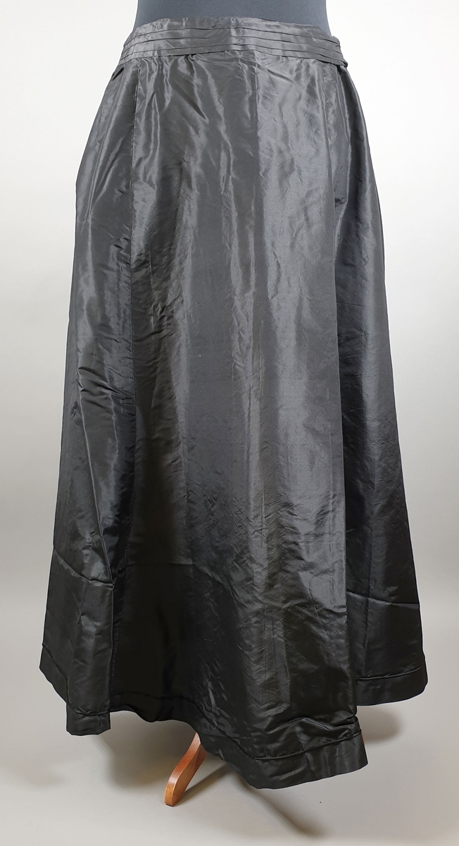 Kjole bestående av to deler - kjoleliv og skjørt. Kjolelivet har dobbelt stoff over rygg og byste, med skjortekrage og slag. Lukkes foran med trykknapper under pynteknapper på det underste laget. Fôret med lin. Svart skjørt med sløyfe på ryggen. Lukkes i ryggen med trykknapper. Stor vidde på skjørtet.