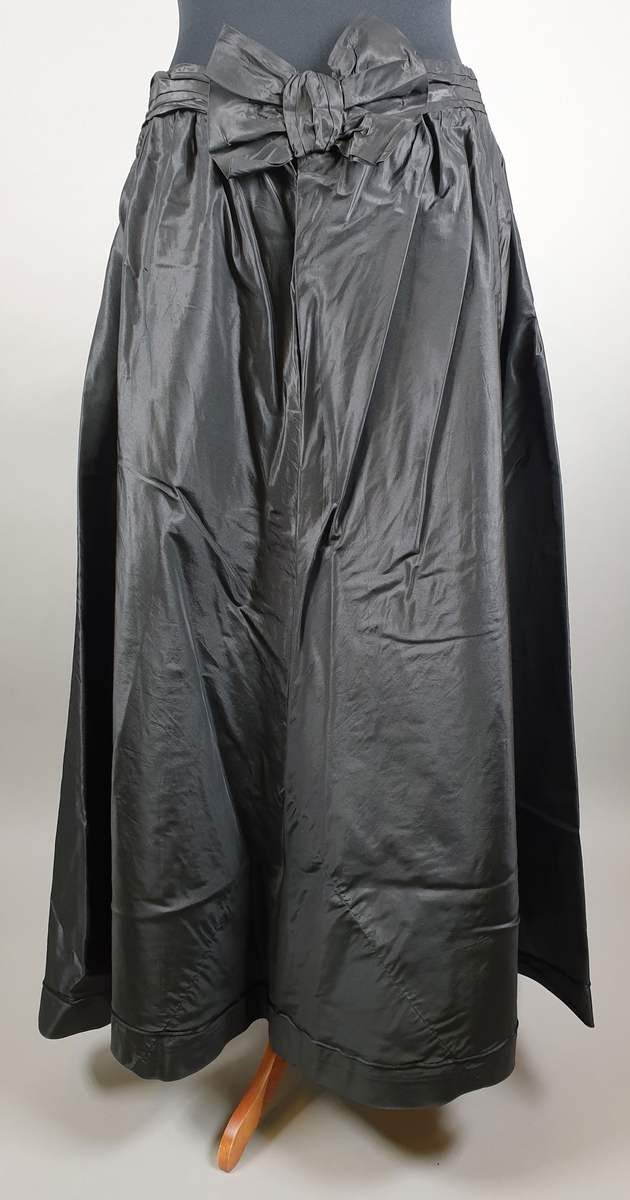 Kjole bestående av to deler - kjoleliv og skjørt. Kjolelivet har dobbelt stoff over rygg og byste, med skjortekrage og slag. Lukkes foran med trykknapper under pynteknapper på det underste laget. Fôret med lin. Svart skjørt med sløyfe på ryggen. Lukkes i ryggen med trykknapper. Stor vidde på skjørtet.