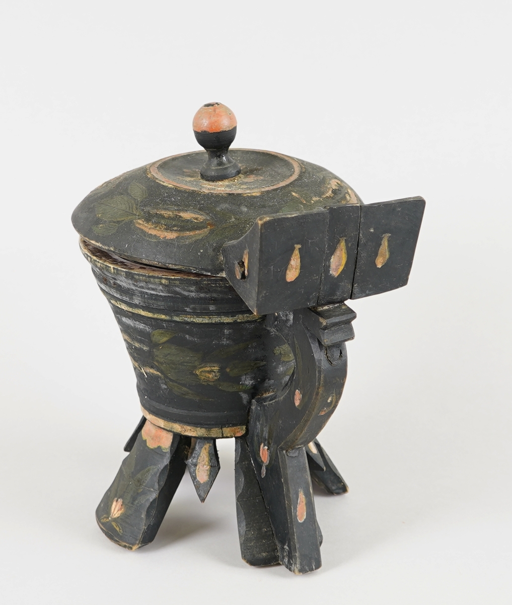 Dreia smørkanne, med påsett lok og hank. Fire føt. Heilt i tre. Korpus har rundt tverrsnitt, og utskrånande sider oppover. Loket er hengsla med ein trenagl øvst i hanken. Innvendig er loket dekorert med utskore mønster. Heile utsida er dekormåla. Føtene er skore ut i same emne som kanna, samt fire "tappar" mellom føtene - til dekor. Laus botnplate ligg inni.