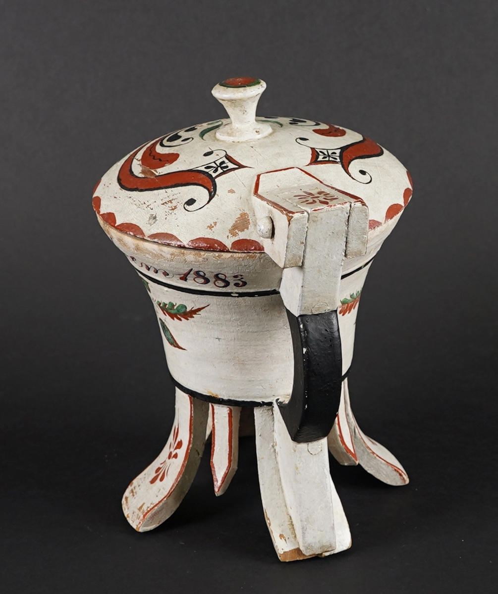 Dreia smørkanne, med påsett lok og hank. Fire føt. Heilt i tre. Korpus har rundt tverrsnitt, og utskrånande sider oppover. Loket er hengsla med ein trenagl øvst i hanken. Innvendig er loket dekorert med utskore mønster. Heile utsida er dekormåla. Føtene er skore ut i same emne som kanna, samt fire "tappar" mellom føtene - til dekor. Gjennomgåande hol i botn, for å hindre oppsprekking ved tørking.