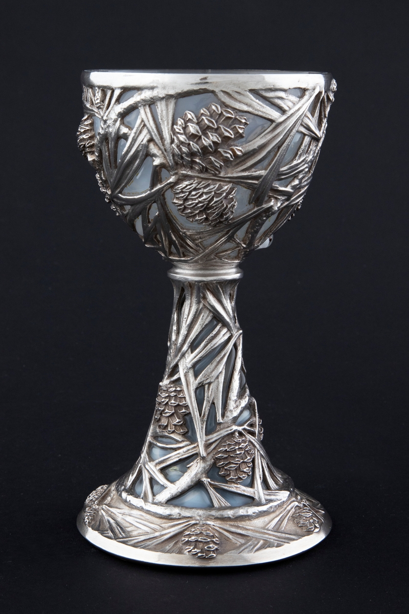 Pokal utført i støpt sølv og halvgjennomskinnelig melkehvitt opalinglass. Det gjennombrutte sølvet danner et flettverk av pinjekongler, grener og nåler i relieff, som brer seg fra den halvkuleformede kupaen og nedover den koniske stetten. Overgangen mellom disse to delene er markert med en ringformet vulst. Inne i pokalen er det innblåst opalinglass, som delvis trer frem som glassblærer på dens ytterside. På foten er det rektangulære felter med stiliserte kongler og grener i lavt relieff.