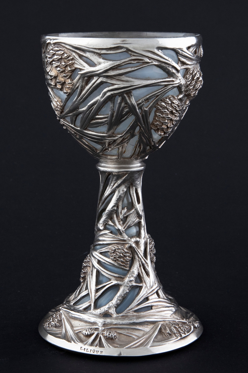 Pokal utført i støpt sølv og halvgjennomskinnelig melkehvitt opalinglass. Det gjennombrutte sølvet danner et flettverk av pinjekongler, grener og nåler i relieff, som brer seg fra den halvkuleformede kupaen og nedover den koniske stetten. Overgangen mellom disse to delene er markert med en ringformet vulst. Inne i pokalen er det innblåst opalinglass, som delvis trer frem som glassblærer på dens ytterside. På foten er det rektangulære felter med stiliserte kongler og grener i lavt relieff.