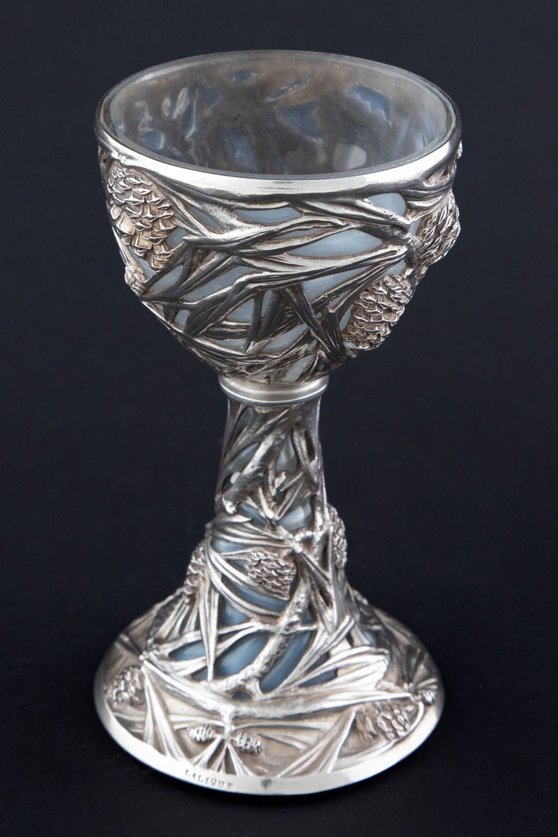 Pokal utført i støpt sølv og halvgjennomskinnelig melkehvitt opalinglass. Det gjennombrutte sølvet danner et flettverk av pinjekongler, grener og nåler i relieff, som brer seg fra den halvkuleformede kupaen og nedover den koniske stetten. Overgangen mellom disse to delene er markert med en ringformet vulst. Inne i pokalen er det innblåst opalinglass, som delvis trer frem som glassblærer på dens ytterside. På foten er det rektangulære felter med stiliserte kongler og grener i lavt relieff.
