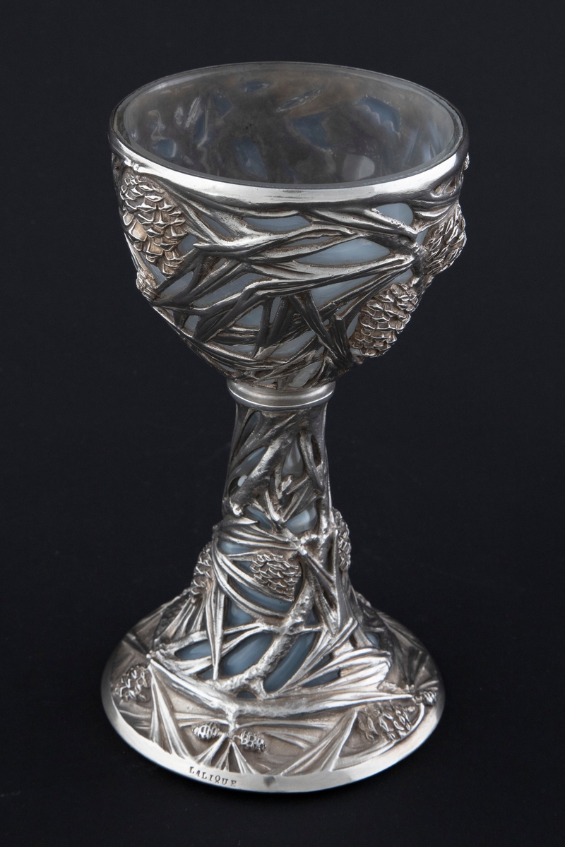 Pokal utført i støpt sølv og halvgjennomskinnelig melkehvitt opalinglass. Det gjennombrutte sølvet danner et flettverk av pinjekongler, grener og nåler i relieff, som brer seg fra den halvkuleformede kupaen og nedover den koniske stetten. Overgangen mellom disse to delene er markert med en ringformet vulst. Inne i pokalen er det innblåst opalinglass, som delvis trer frem som glassblærer på dens ytterside. På foten er det rektangulære felter med stiliserte kongler og grener i lavt relieff.