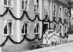 Elva kvinnor i långa ljusa sommarklänningar och en man i ljust och hatt står på trappan till prästgården vid Brahegatan i Gränna. Kvinnorna har blomsterbuketter i händerna. Byggnaden är dekorerard med blomstergirlanger. Man väntar på kronprinsparet Gustaf och Margaret.