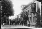 En grupp med barn och vuxna på Brahegatan utanför Hjertströmska gården som är dekorerad med blomstergirlanger och unionsflaggor. De väntar på kung Oscar II. Flaggstång med unionsflagg i gathörnet.