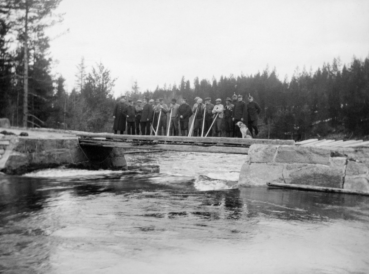 Dam med fløtermannskap og statspoliti i Julussvassdraget i Elverum.  Fotografiet er tatt fra dammen mot tømmerløpet, hvor vannet strømmer fritt mot det nedenforliggende vassdraget.  Dammen er en murkonstruksjon av tuktet gråstein. På dambrua står 16 karer - fløtere og konstabler fra statspolitiet. Politioppbudet skyldtes den tilspissete situasjonen under fløtinga i dette vassdraget i mai 1927. Åstyret hadde gitt Hans Mellum fløterhusbondstatusen i konkurranse med Martin Halstensens lag, som besto av folk med sympatier for fagorganisasjonen. Dette utløste blokade med til dels væpnete demonstranter, noe som fikk Justisdepartementet til å sende statspoliti med hunder til Julussa for å beskytte Mellums fløterlag.