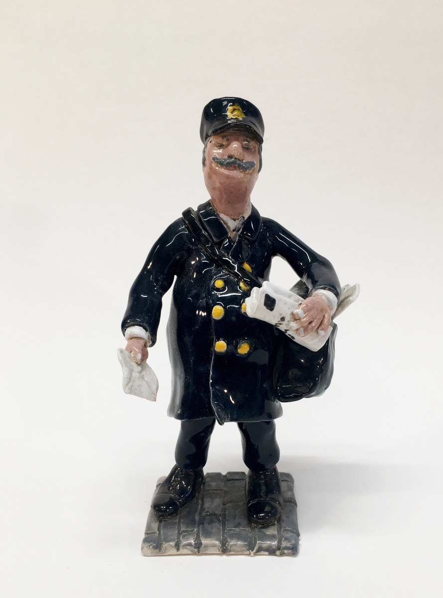 Figurin förställande en brevbärare med uniform, väska och mössa. Han håller ett brev i handeln.