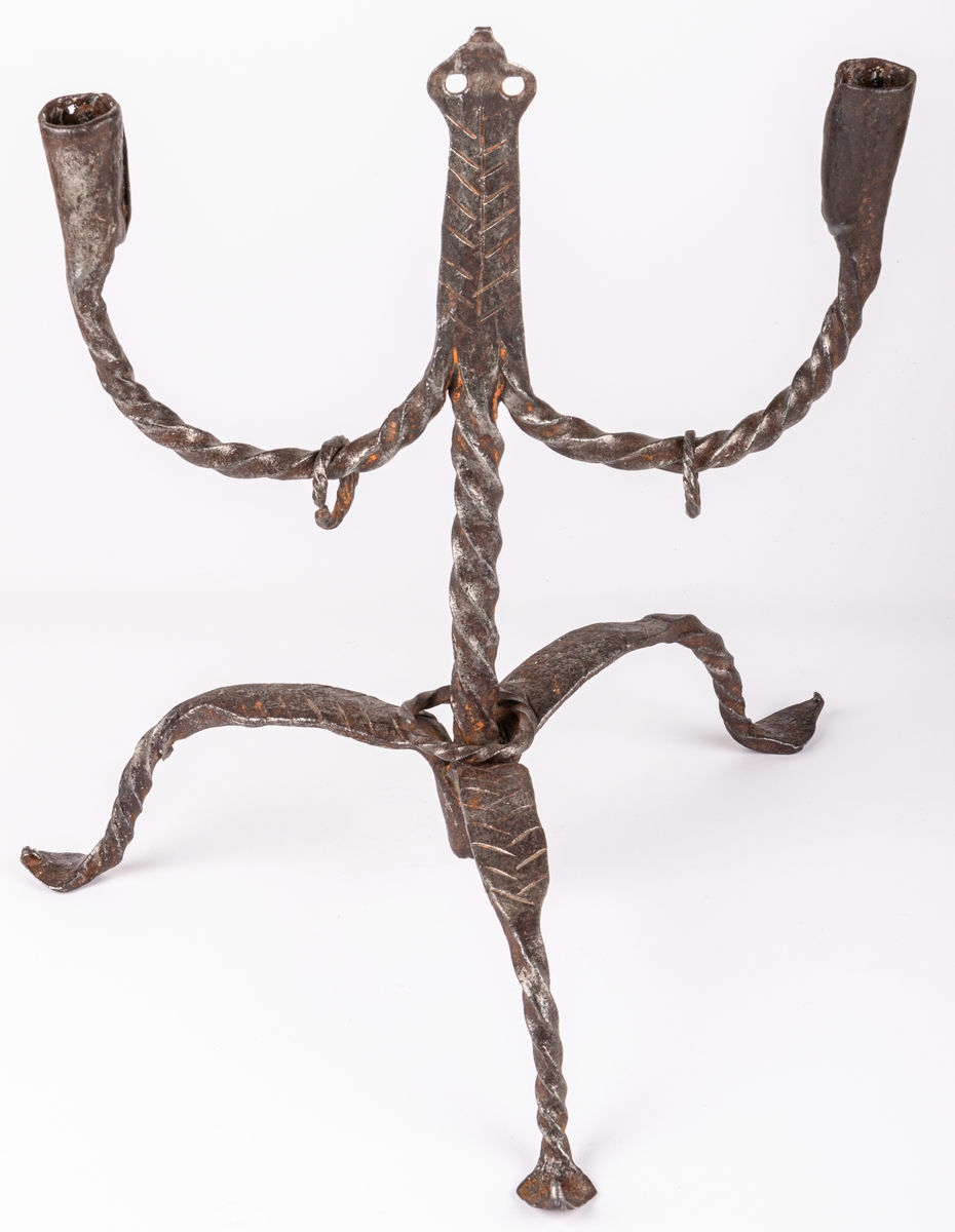 Tvåarmad ljusstake av järn. 3 fötter. Snodda tenar. I mitten en ten sluttande i en rund platta

Lappkort. Tre bågböjda fötter, två ljusarmar med smidda holkar. I mitten trebladig uppståndare, med två små hål där det troligen hängt löv eller liknande prydnad. Järntenarna spiralvridna.