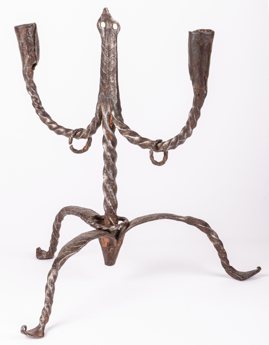 Tvåarmad ljusstake av järn. 3 fötter. Snodda tenar. I mitten en ten sluttande i en rund platta

Lappkort. Tre bågböjda fötter, två ljusarmar med smidda holkar. I mitten trebladig uppståndare, med två små hål där det troligen hängt löv eller liknande prydnad. Järntenarna spiralvridna.