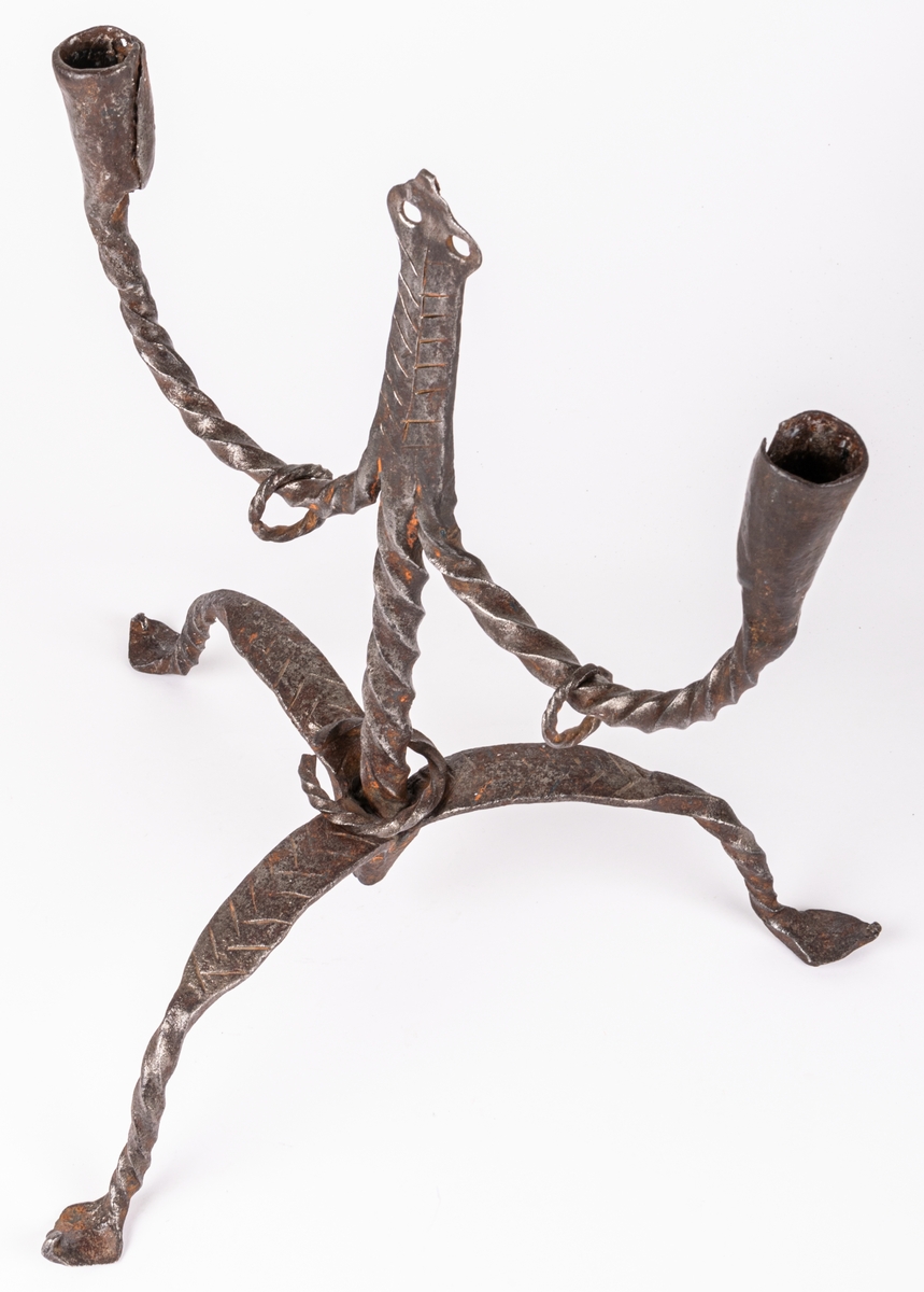 Tvåarmad ljusstake av järn. 3 fötter. Snodda tenar. I mitten en ten sluttande i en rund platta

Lappkort. Tre bågböjda fötter, två ljusarmar med smidda holkar. I mitten trebladig uppståndare, med två små hål där det troligen hängt löv eller liknande prydnad. Järntenarna spiralvridna.