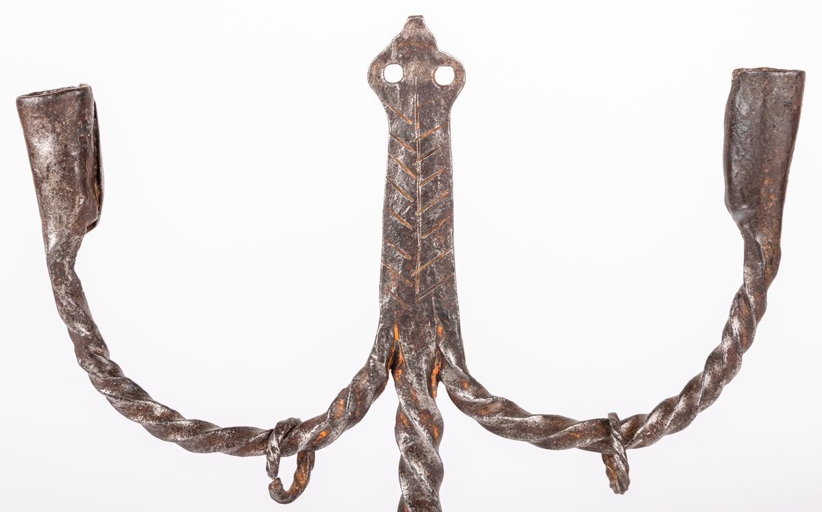 Tvåarmad ljusstake av järn. 3 fötter. Snodda tenar. I mitten en ten sluttande i en rund platta

Lappkort. Tre bågböjda fötter, två ljusarmar med smidda holkar. I mitten trebladig uppståndare, med två små hål där det troligen hängt löv eller liknande prydnad. Järntenarna spiralvridna.