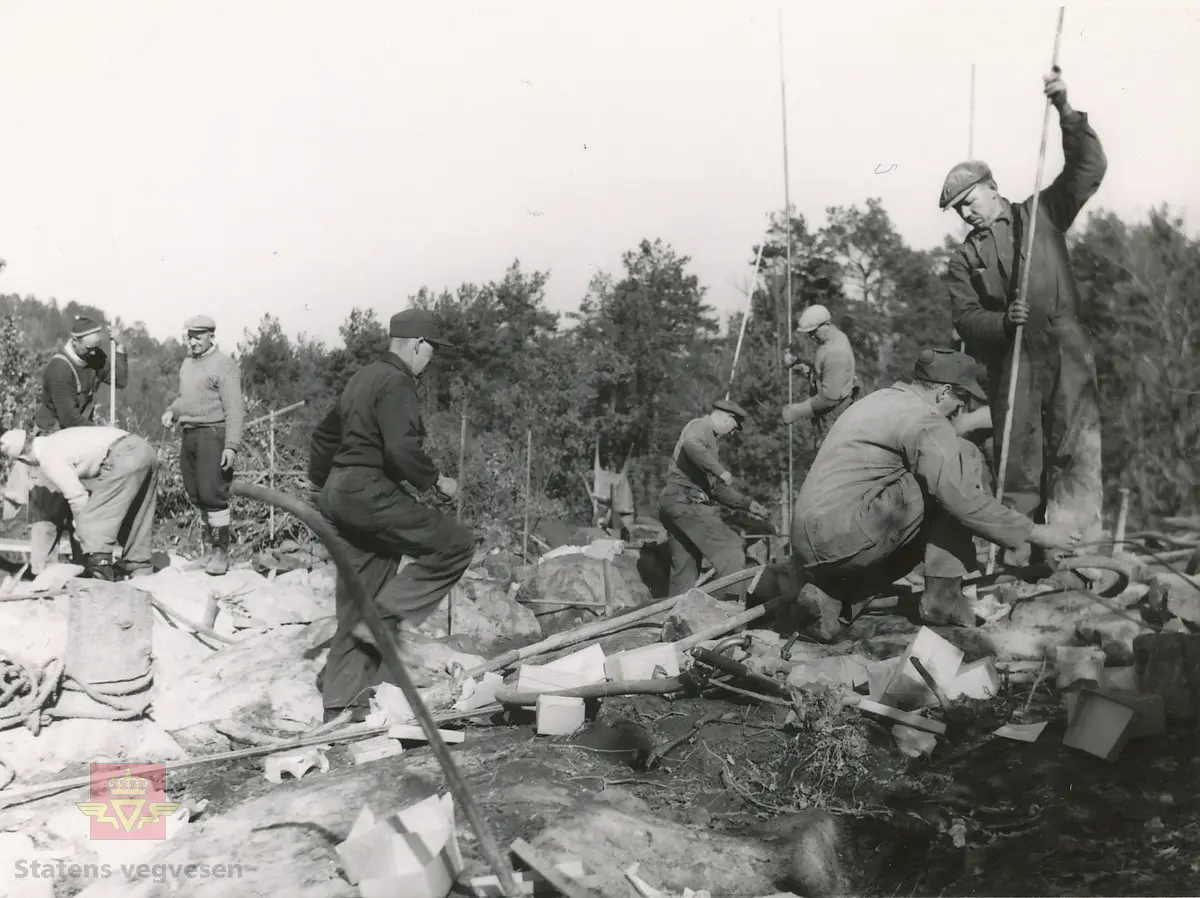 Bygging av Tromøybrua 1959-1961. 
Mange er i sving for å gjøre klart til sprengning. Skytebasen  svinger ladekjeppen. Trestikkene i bakgrunnen er stikningsgrunnlaget for boringen. Den med tverrstikk gir informasjon om retning, hvor dypt det skal bores m.m. Proppen tas ut av borehullene, stappes med sprengstoff: tenneren i bunnen, tilkoblet den elektriske ledning, deretter dynamitt, glynitt og fordemming med sand i toppen så energien ikke skal forsvinne opp, men til sidene. 

Jf. bilder NVM 09-F-00331-00364 og NVM 09-F-00722-00744.