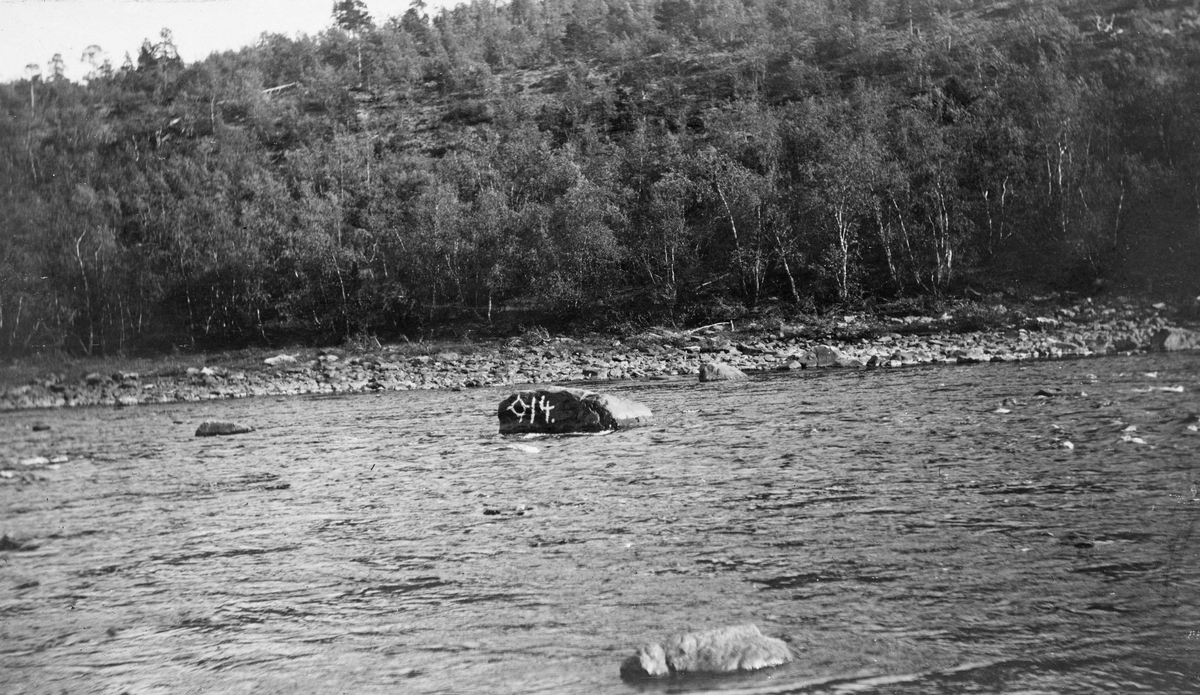 Elvelandskap ved «Mundfossen» i Karasjok i Finnmark, antakelig i 1913. Fotografiet er tatt med kameraet vendt på tvers av elva, som er forholdsvis bred og grunn. I elveløpet stikker det opp en del steinbloker. Den største av dem var påmalt tallet «14» med kvit farge da dette fotografiet ble tatt. Dette hadde sammenheng med at denne steinen skulle sprenges. Det hadde nemlig vist seg at fløtingstømmeret fra statsskogene i området hadde en tendens til å hekte seg fast og vase seg sammen ved denne og 13 andre steiner i dette elveløpet. Derfor leide skogforvalteren i området mannskap som skulle sprenge de merkede steinene vinteren 1913-14.