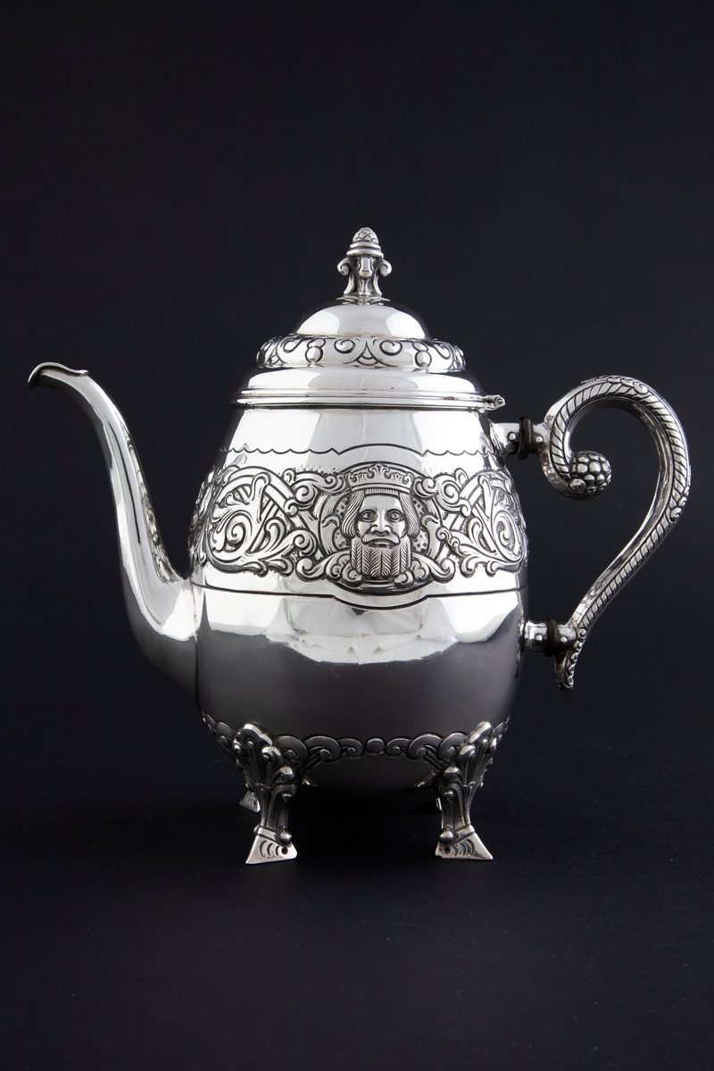 Kaffekanne med ovalformet korpus, som hviler på fire ben. Rundt midten av korpus løper en billedfrise bestående av motivscener, masker og flettverkornamentikk. Hengslet sirkulært lokk, hvor nedre del er dekorert med drevet og siselert flettverksornamentikk. Øvre del er hjelmformet med profilert lokknapp. Spiralformet hank som løper fra nedre del av korpus til skulderen. Svungen tut.