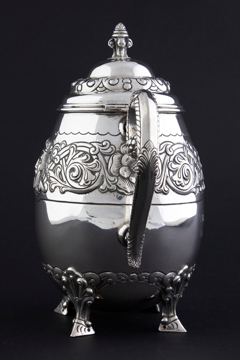 Kaffekanne med ovalformet korpus, som hviler på fire ben. Rundt midten av korpus løper en billedfrise bestående av motivscener, masker og flettverkornamentikk. Hengslet sirkulært lokk, hvor nedre del er dekorert med drevet og siselert flettverksornamentikk. Øvre del er hjelmformet med profilert lokknapp. Spiralformet hank som løper fra nedre del av korpus til skulderen. Svungen tut.