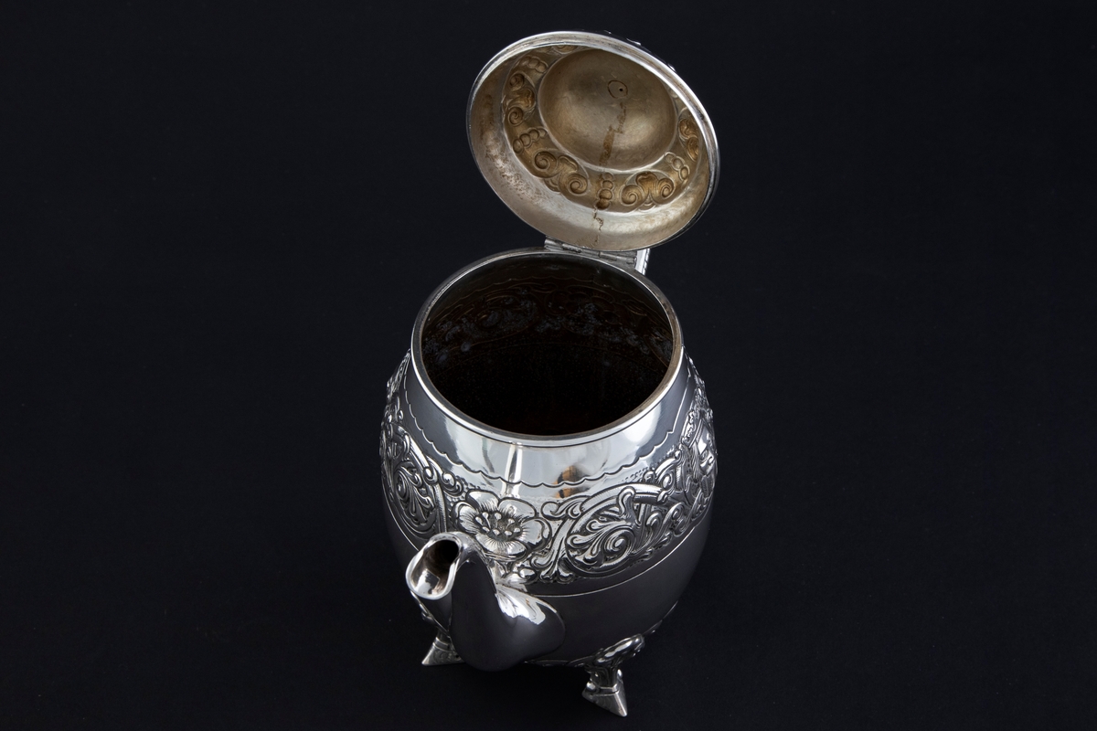 Kaffekanne med ovalformet korpus, som hviler på fire ben. Rundt midten av korpus løper en billedfrise bestående av motivscener, masker og flettverkornamentikk. Hengslet sirkulært lokk, hvor nedre del er dekorert med drevet og siselert flettverksornamentikk. Øvre del er hjelmformet med profilert lokknapp. Spiralformet hank som løper fra nedre del av korpus til skulderen. Svungen tut.