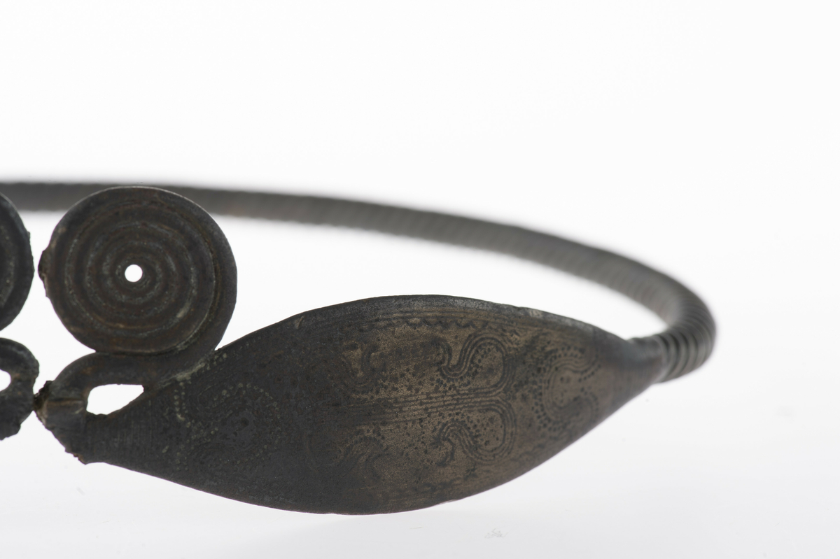 Vriden ring med ovala, böljebandsdekorerade ändplattor som avslutas med spiralupprullningar. Av i hopfogningen mitt emellan spiralupprullningarna, spricka i ringen på motsatta sidan.
