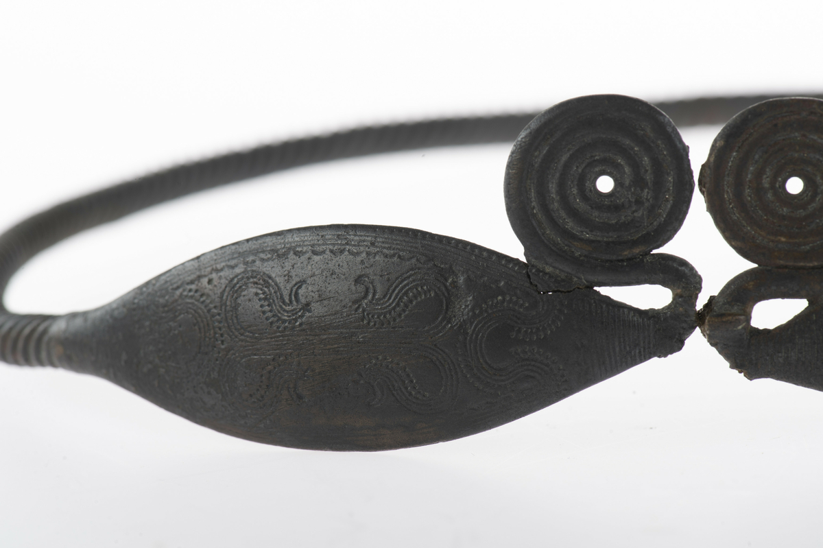 Vriden ring med ovala, böljebandsdekorerade ändplattor som avslutas med spiralupprullningar. Av i hopfogningen mitt emellan spiralupprullningarna, spricka i ringen på motsatta sidan.