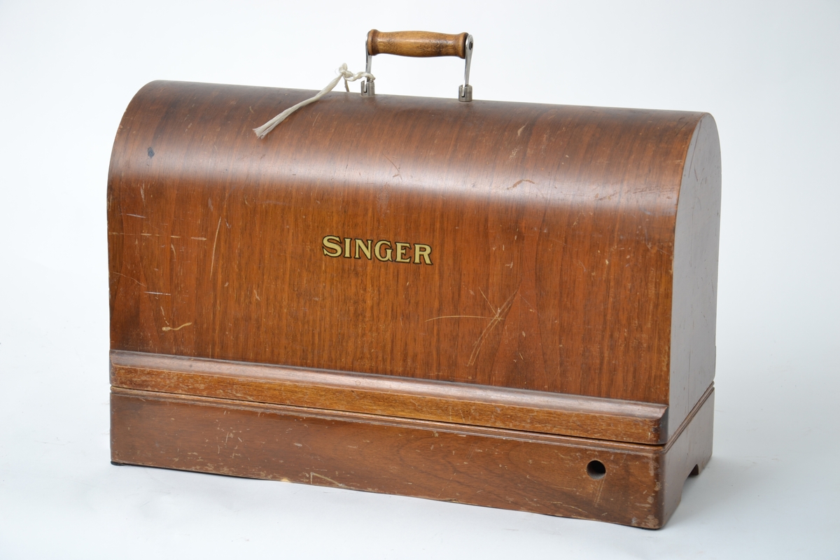 Elektrisk symaskin med fotpedal. Maskinen har et futteral i tre med nøkler. Håndtak på toppen av lokket. Maskinen kan vippes opp fra konsollen den sitter på.