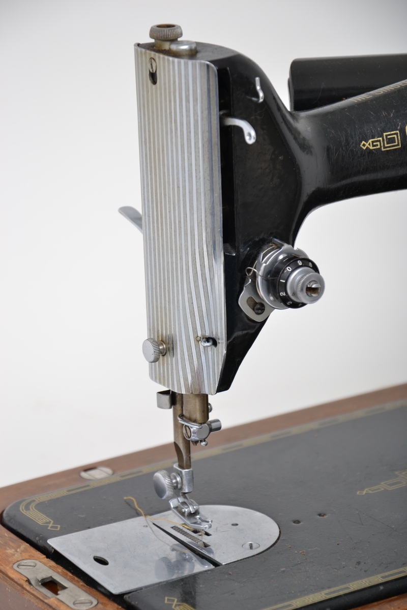 Elektrisk symaskin med fotpedal. Maskinen har et futteral i tre med nøkler. Håndtak på toppen av lokket. Maskinen kan vippes opp fra konsollen den sitter på.