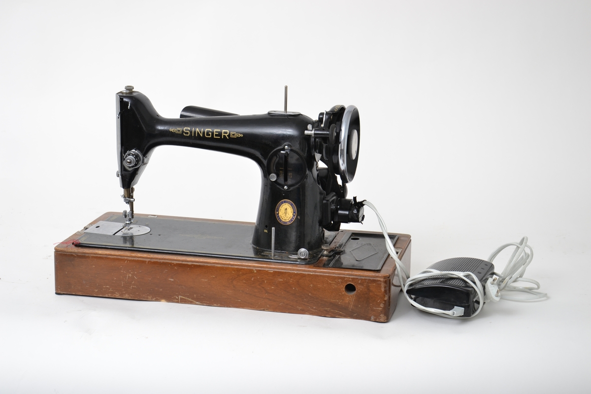 Elektrisk symaskin med fotpedal. Maskinen har et futteral i tre med nøkler. Håndtak på toppen av lokket. Maskinen kan vippes opp fra konsollen den sitter på.