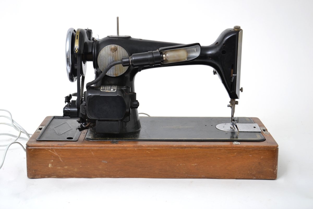 Elektrisk symaskin med fotpedal. Maskinen har et futteral i tre med nøkler. Håndtak på toppen av lokket. Maskinen kan vippes opp fra konsollen den sitter på.