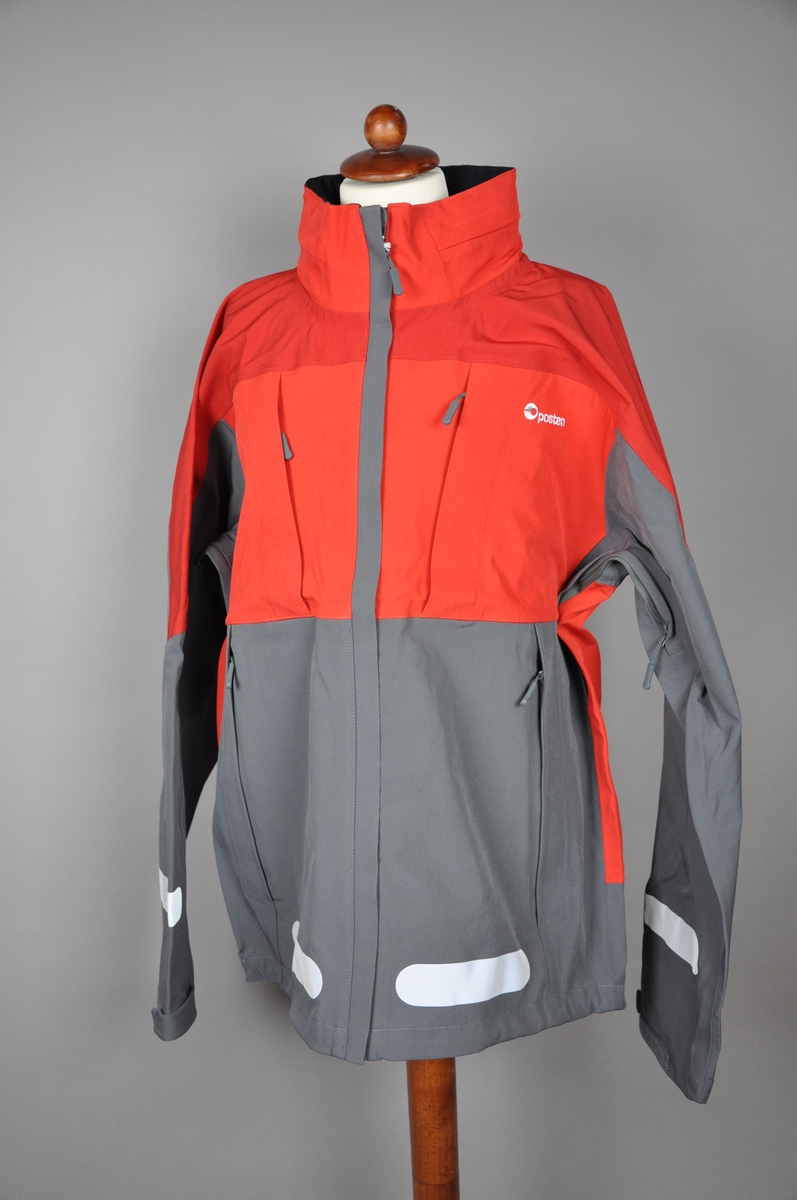 Rød og grå skalljakke. Postens logo på ryggen og et mindre brodert logomerke på fremsiden. Jakken er størrelse Large i herremodell. Innsiden er foret med tynt polyester fleece. 

Reflekser på fram- og bakside og på ermer. Jakken har to brystlommer, den ene har en foret mobillomme på innsiden. To store side lommer, og åpning med glidelås under ermene. I kragen er det festet en hette, den ligger i en innsydd lomme i kragen og kan tas ut ved behov.