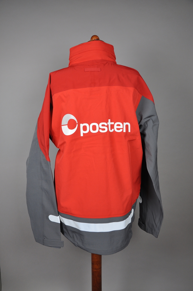 Rød og grå skalljakke. Postens logo på ryggen og et mindre brodert logomerke på fremsiden. Jakken er størrelse Large i herremodell. Innsiden er foret med tynt polyester fleece. 

Reflekser på fram- og bakside og på ermer. Jakken har to brystlommer, den ene har en foret mobillomme på innsiden. To store side lommer, og åpning med glidelås under ermene. I kragen er det festet en hette, den ligger i en innsydd lomme i kragen og kan tas ut ved behov.