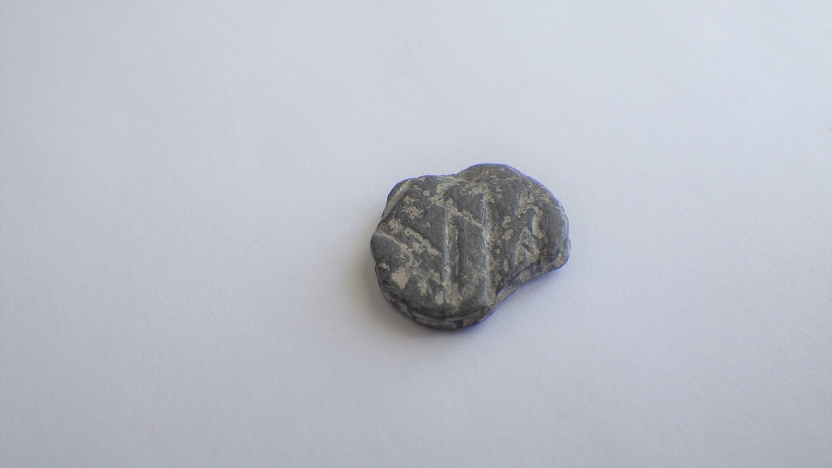 Begge sider viser Konturer av et våpenskjold. den ene siden viser et mulig skrått bånd fra øverst til venstre og ned til høyre som deler skjoldet. den samme plaketten har svake antydninger til kroppen av et stående fabeldyr til høyre for skjoldet. 