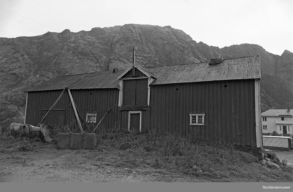 Bygninger i fiskeværet Nusfjord. Fjøs med langveggen imot. Liten ark med skyvedør. 2 dører i 1. etg og 2 små vinduer. Fjell i bakgrunnen.