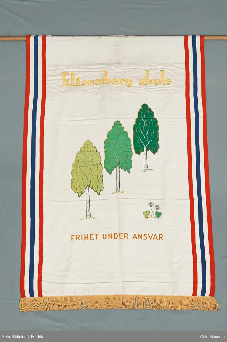 Fløtefarget silke med applikasjonsbroderi. Dobbel duk. På begge siders langsider to røde og en blå langsgående stripe. På midten tre bjerketrær i nyanser av grønt med grener og stammer i sjattersøm. Blåveis. Over trærne i gult: Elisenberg skole. Under trærne i gul sjattersøm: Frihet under ansvar. På baksiden i sjattersøm i gult: V P S 1898. Nedkanten med gul silkefrynse.