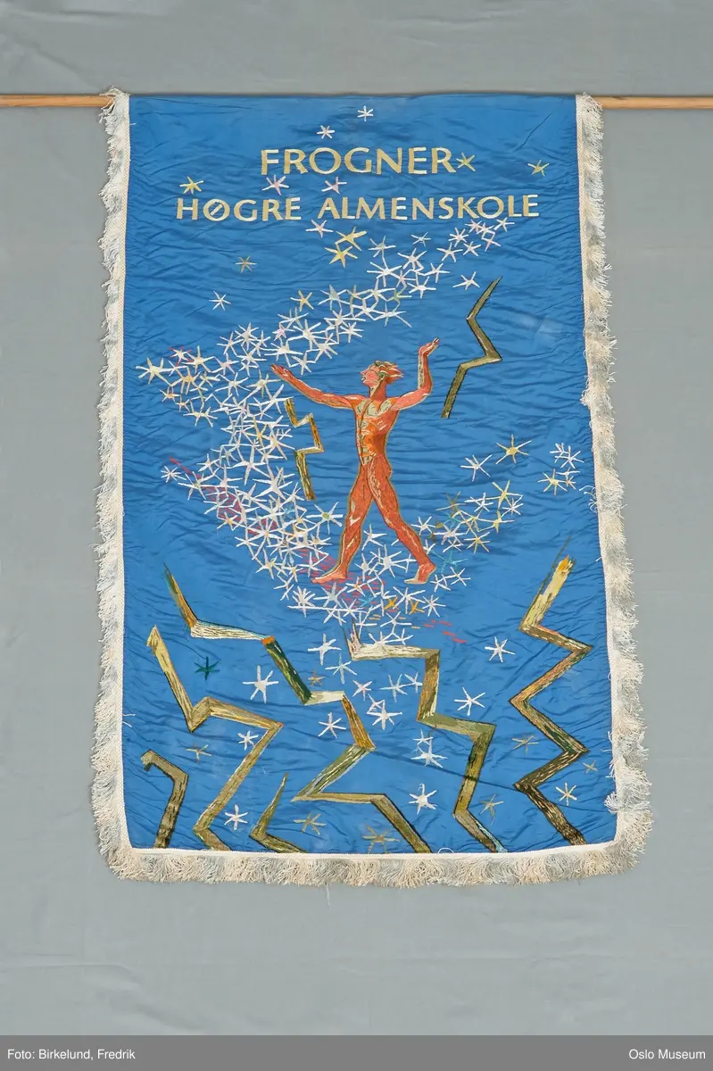 Duk av kongeblå dobbel silke kantet med hvit silkeagraman m. frynser. En side med broderi i sjattersøm i polykromi: øverst med gule bokstaver: Frogner Høgre Almenskole. Under dette stjernehimmel med hvite stjerner og lyn? i grønt/ gult, på midten mannsskikkelse i rødt/ grønt/ beige.