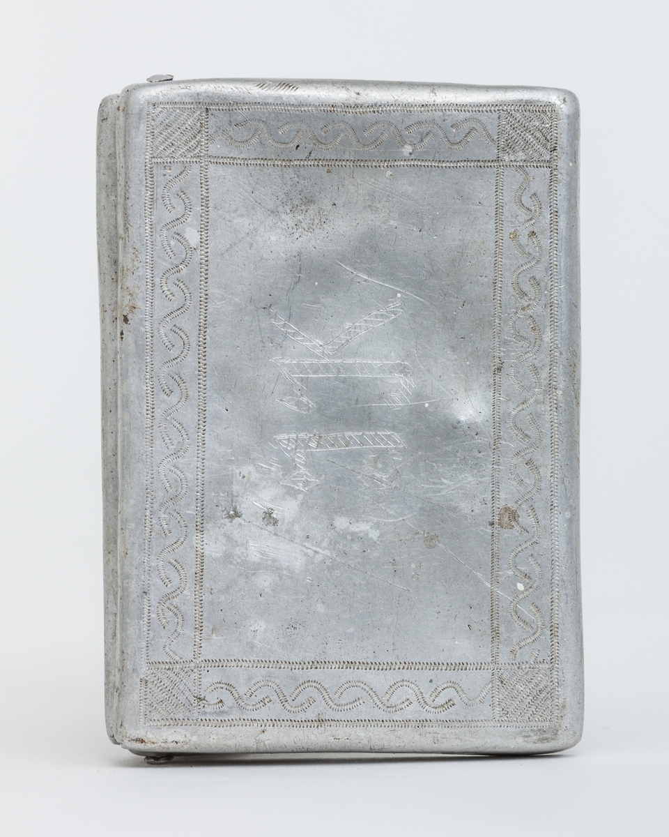 Sigarettetui lagert av sovjetiske krigsfange. Framstil av aluminium og lokket er dekorert med stempelpregede border og initialene I (J?)K. Lokket er klinket fast med en liten bolt i hver kortende.