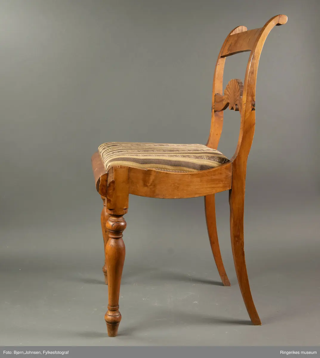 Biedermeierstol i lyst treverk. Forbeina er dreid, med ringer og skiver. Sargen foran på stolen har en buet profil. Setet er trapesformet. Side-sargene er også skåret til med fin profil. Setet er løst. Stolryggen er elegant, ryggbrettet er vannrett, og er pent skåret ut. Utskjæringen minner mest om et skjell, med bølger på hver side. Akterstavene ender i to små runde former. Toppstykket er ikke skåret ut. Stolsetet er en pute, som er trukket med stoff med svarte og gule striper. (Flere steder er den svarte fargen bleknet, og ser nå mere brun ut). De gule stripene har innslag av gull i geometriske mønster. Noen svarte striper er smale og ikke dekorerte. Andre svarte striper har blomsterdekorasjon, de smaleste med rosa og gule småblomster og grønne blad, og de bredere stripene med kranser og kryss med rosa blomster, men også med innslag av gule og blå blomster, samt grønne blader.