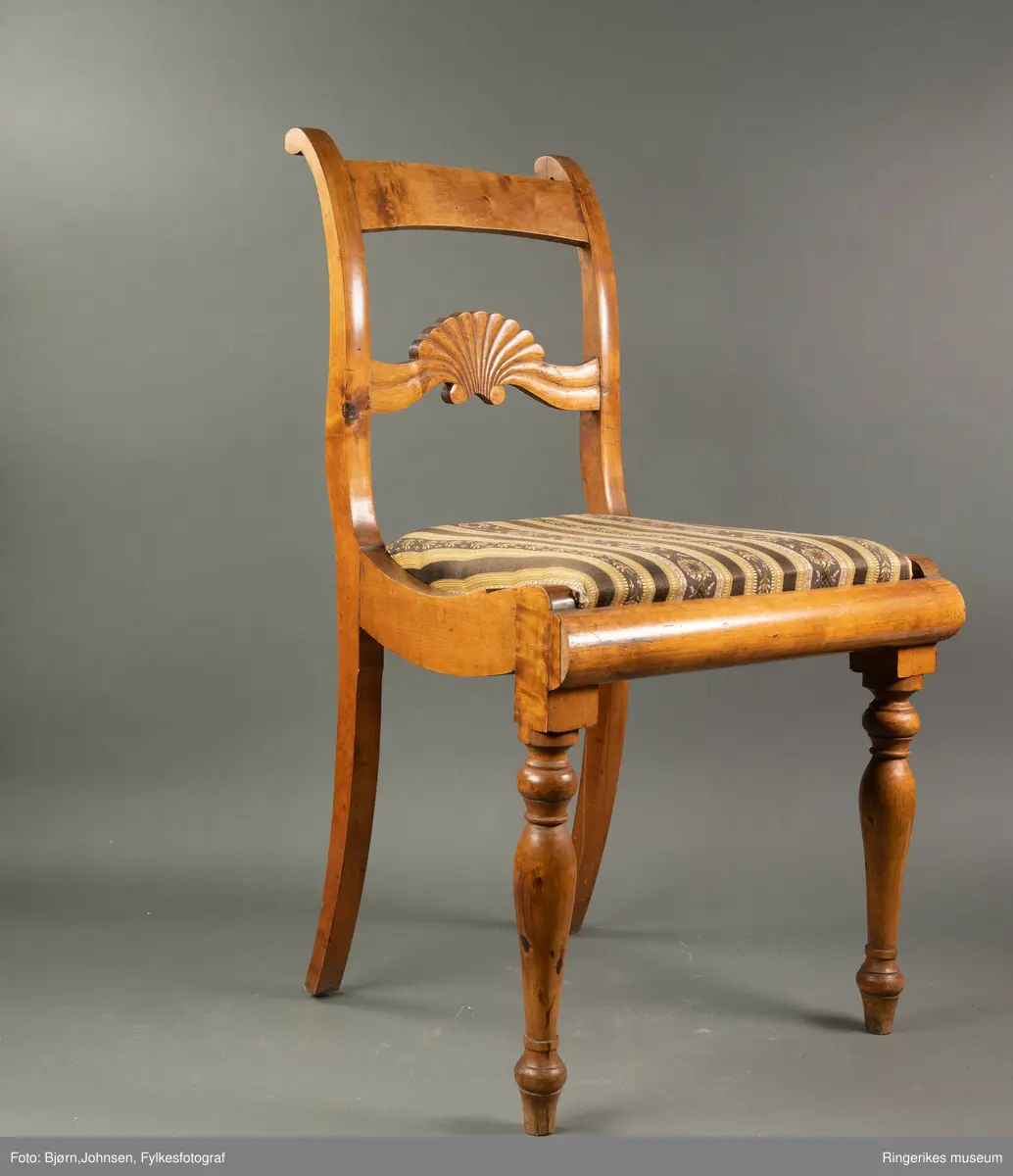 Biedermeierstol i lyst treverk. Forbeina er dreid, med ringer og skiver. Sargen foran på stolen har en buet profil. Setet er trapesformet. Side-sargene er også skåret til med fin profil. Setet er løst. Stolryggen er elegant, ryggbrettet er vannrett, og er pent skåret ut. Utskjæringen minner mest om et skjell, med bølger på hver side. Akterstavene ender i to små runde former. Toppstykket er ikke skåret ut. Stolsetet er en pute, som er trukket med stoff med svarte og gule striper. (Flere steder er den svarte fargen bleknet, og ser nå mere brun ut). De gule stripene har innslag av gull i geometriske mønster. Noen svarte striper er smale og ikke dekorerte. Andre svarte striper har blomsterdekorasjon, de smaleste med rosa og gule småblomster og grønne blad, og de bredere stripene med kranser og kryss med rosa blomster, men også med innslag av gule og blå blomster, samt grønne blader.