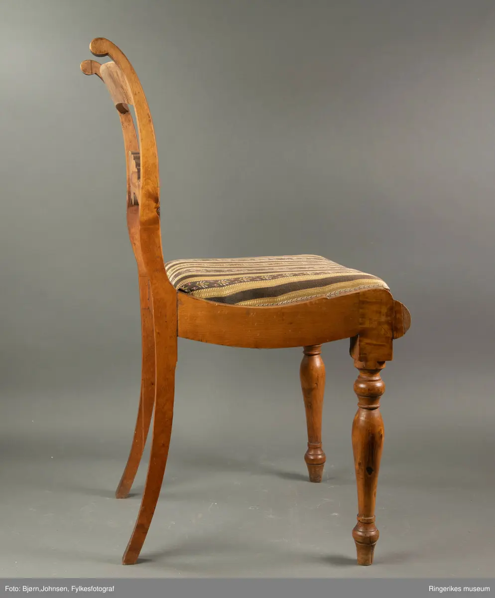 Biedermeierstol i lyst treverk. Forbeina er dreid, med ringer og skiver. Sargen foran på stolen har en buet profil. Setet er trapesformet. Side-sargene er også skåret til med fin profil. Setet er løst. Stolryggen er elegant, ryggbrettet er vannrett, og er pent skåret ut. Utskjæringen minner mest om et skjell, med bølger på hver side. Akterstavene ender i to små runde former. Toppstykket er ikke skåret ut. Stolsetet er en pute, som er trukket med stoff med svarte og gule striper. (Flere steder er den svarte fargen bleknet, og ser nå mere brun ut). De gule stripene har innslag av gull i geometriske mønster. Noen svarte striper er smale og ikke dekorerte. Andre svarte striper har blomsterdekorasjon, de smaleste med rosa og gule småblomster og grønne blad, og de bredere stripene med kranser og kryss med rosa blomster, men også med innslag av gule og blå blomster, samt grønne blader.
