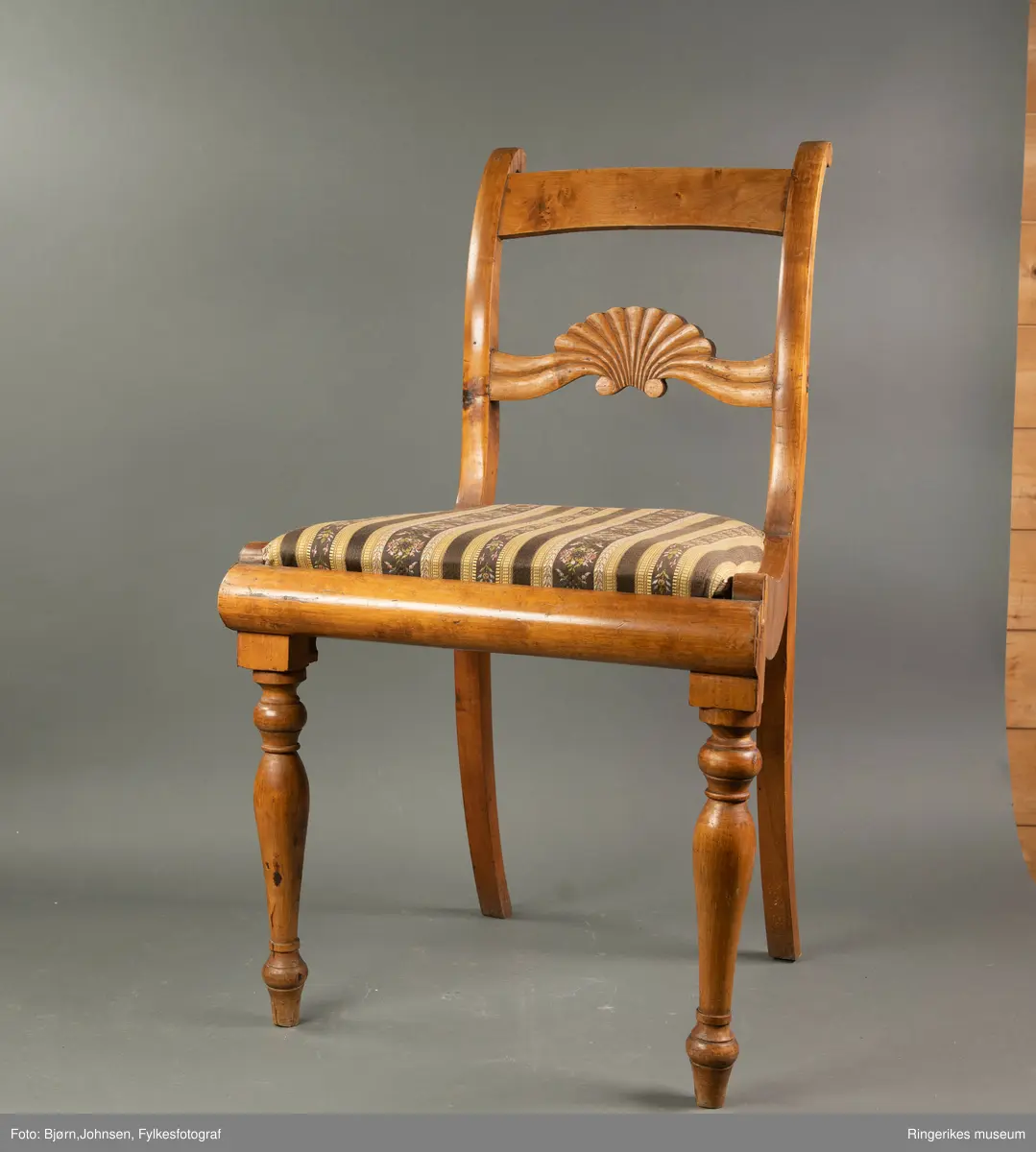Biedermeierstol i lyst treverk. Forbeina er dreid, med ringer og skiver. Sargen foran på stolen har en buet profil. Setet er trapesformet. Side-sargene er også skåret til med fin profil. Setet er løst. Stolryggen er elegant, ryggbrettet er vannrett, og er pent skåret ut. Utskjæringen minner mest om et skjell, med bølger på hver side. Akterstavene ender i to små runde former. Toppstykket er ikke skåret ut. Stolsetet er en pute, som er trukket med stoff med svarte og gule striper. (Flere steder er den svarte fargen bleknet, og ser nå mere brun ut). De gule stripene har innslag av gull i geometriske mønster. Noen svarte striper er smale og ikke dekorerte. Andre svarte striper har blomsterdekorasjon, de smaleste med rosa og gule småblomster og grønne blad, og de bredere stripene med kranser og kryss med rosa blomster, men også med innslag av gule og blå blomster, samt grønne blader.