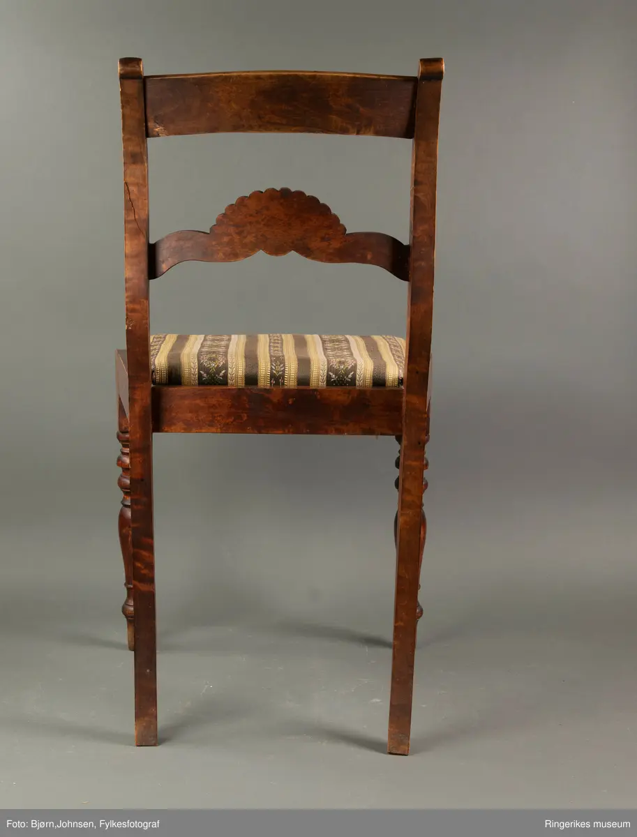 Biedermeierstol i mørkt treverk. Forbeina er dreid i markerte ringer og skiver. Bakbeina er ikke forseggjort. Setet er trapesformet, og sitter løst. Sargen foran har en buet profil. Ryggbrettet er vannrett, og er formet som et skjell med bølger foran. Akterstavene er skåret ut til runde former på toppen. Putene er trukket med stoff med svarte og gule striper. De gule stripene har innslag av gull i geometriske former. Noen av de svarte stripene er ikke dekorerte. De smaleste av de dekorerte svarte stripene har små blomster i rosa og gult, og grønne blader. De bredeste svarte stripene er dekorert med kranser og kryss, med blomster i rosa, gult og blått, samt grønne blader.