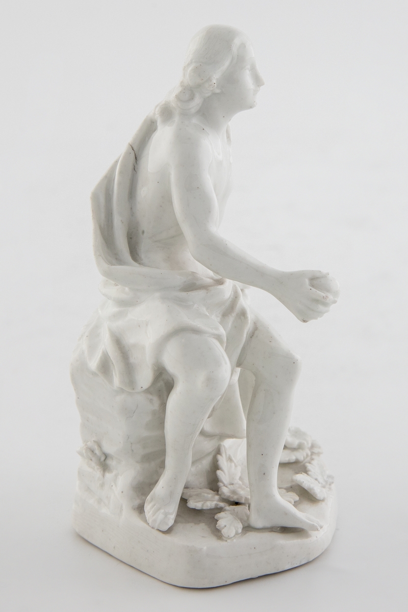 Hvitglasert, umalt porselensfigur med en ung mann sittende på en klippe. Figuren holder et eple i høyre hånd og identifiseres som den trojanske prinsen Paris fra gresk mytologi. Sokkelen er dekorert med blomster og blader.