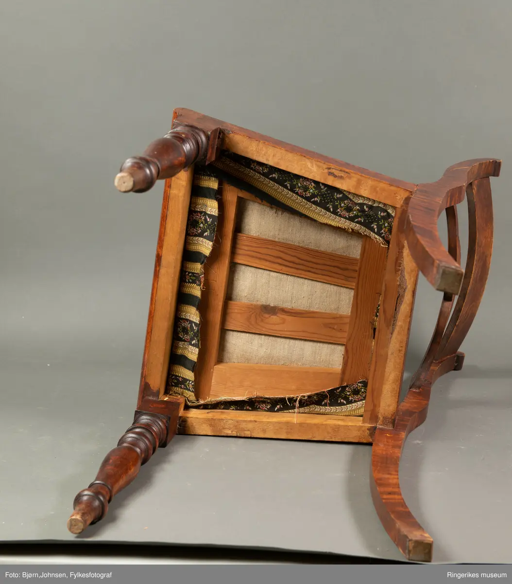 Biedermeierstol i mørkt treverk. Forbeina er dreid i markerte ringer og skiver. Bakbeina er ikke forseggjort. Setet er trapesformet, og sitter løst. Sargen foran har en buet profil. Ryggbrettet er vannrett, og er formet som et skjell med bølger foran. Akterstavene er skåret ut til runde former på toppen. Putene er trukket med stoff med svarte og gule striper. De gule stripene har innslag av gull i geometriske former. Noen av de svarte stripene er ikke dekorerte. De smaleste av de dekorerte svarte stripene har små blomster i rosa og gult, og grønne blader. De bredeste svarte stripene er dekorert med kranser og kryss, med blomster i rosa, gult og blått, samt grønne blader.