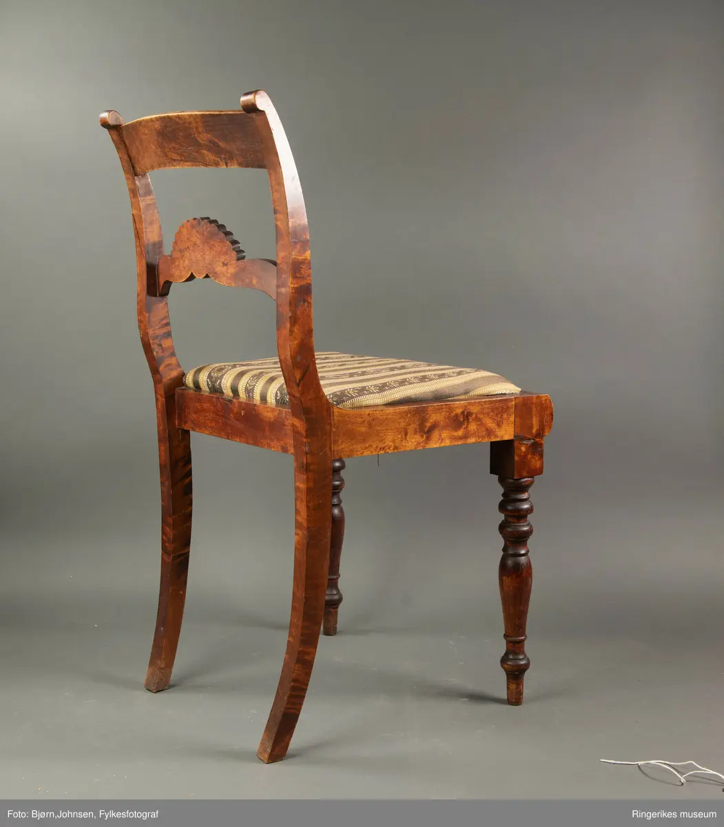 Biedermeierstol i mørkt treverk. Forbeina er dreid i markerte ringer og skiver. Bakbeina er ikke forseggjort. Setet er trapesformet, og sitter løst. Sargen foran har en buet profil. Ryggbrettet er vannrett, og er formet som et skjell med bølger foran. Akterstavene er skåret ut til runde former på toppen. Putene er trukket med stoff med svarte og gule striper. De gule stripene har innslag av gull i geometriske former. Noen av de svarte stripene er ikke dekorerte. De smaleste av de dekorerte svarte stripene har små blomster i rosa og gult, og grønne blader. De bredeste svarte stripene er dekorert med kranser og kryss, med blomster i rosa, gult og blått, samt grønne blader.