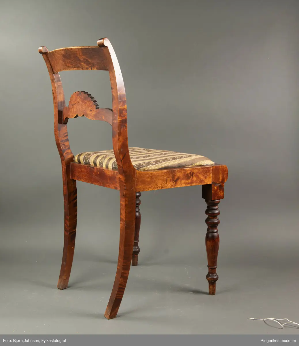 Biedermeierstol i mørkt treverk. Forbeina er dreid i markerte ringer og skiver. Bakbeina er ikke forseggjort. Setet er trapesformet, og sitter løst. Sargen foran har en buet profil. Ryggbrettet er vannrett, og er formet som et skjell med bølger foran. Akterstavene er skåret ut til runde former på toppen. Putene er trukket med stoff med svarte og gule striper. De gule stripene har innslag av gull i geometriske former. Noen av de svarte stripene er ikke dekorerte. De smaleste av de dekorerte svarte stripene har små blomster i rosa og gult, og grønne blader. De bredeste svarte stripene er dekorert med kranser og kryss, med blomster i rosa, gult og blått, samt grønne blader.