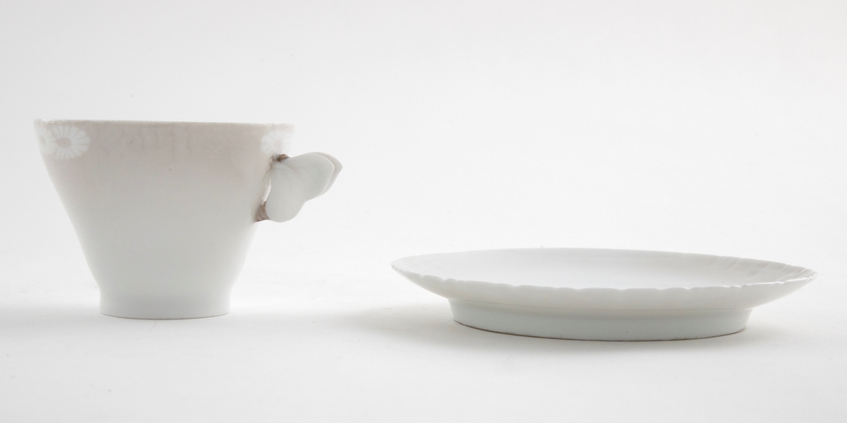 Kaffekopp med margeritter fremstilt langs kanten. Hanken er utformet som en sommerfugl. Tilhørende sirkulært fat. Langs ytterkanten er det fremstilt margeritter samt riller i lavt relieff.