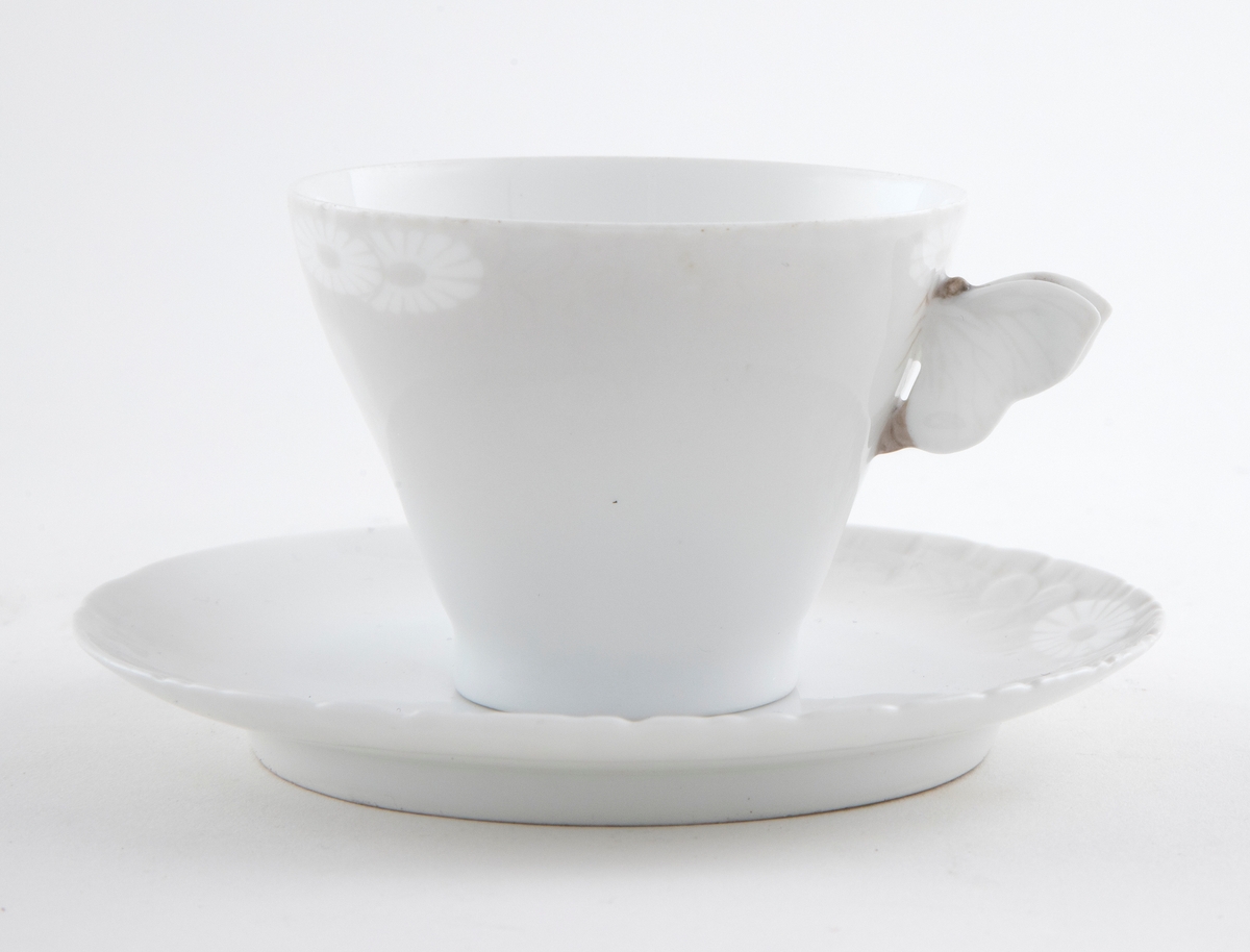 Kaffekopp med margeritter fremstilt langs kanten. Hanken er utformet som en sommerfugl. Tilhørende sirkulært fat. Langs ytterkanten er det fremstilt margeritter samt riller i lavt relieff.