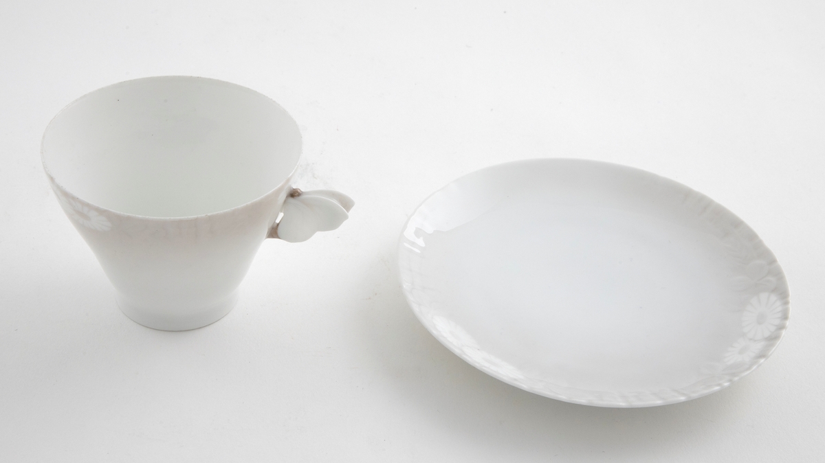 Kaffekopp med margeritter fremstilt langs kanten. Hanken er utformet som en sommerfugl. Tilhørende sirkulært fat. Langs ytterkanten er det fremstilt margeritter samt riller i lavt relieff.