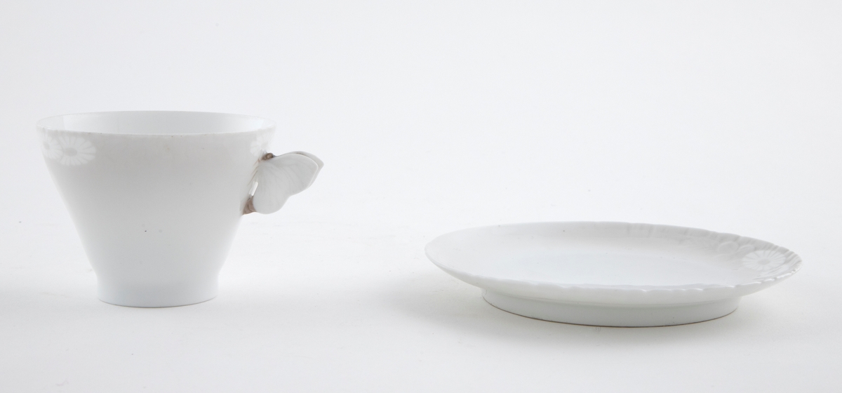 Kaffekopp med margeritter fremstilt langs kanten. Hanken er utformet som en sommerfugl. Tilhørende sirkulært fat. Langs ytterkanten er det fremstilt margeritter samt riller i lavt relieff.