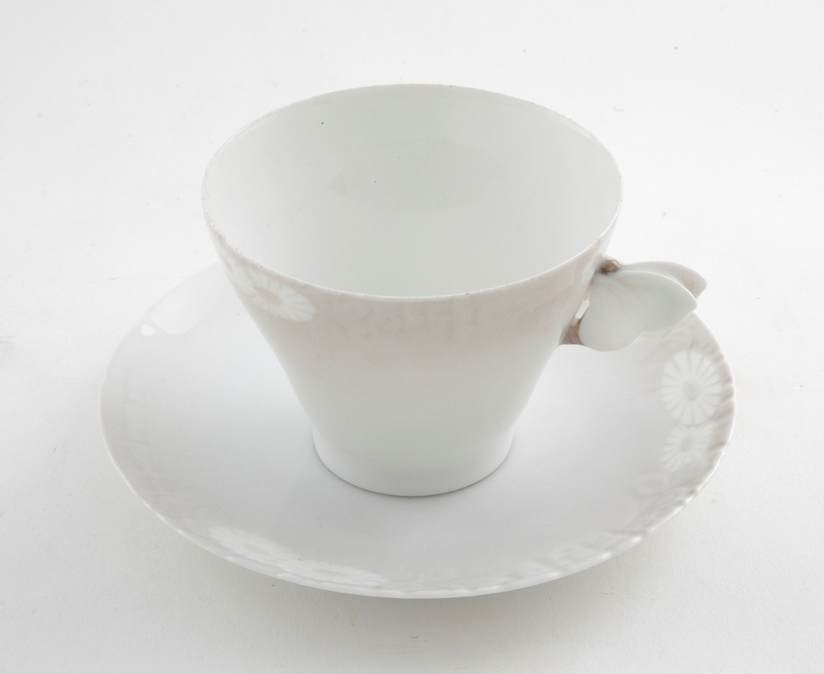 Kaffekopp med margeritter fremstilt langs kanten. Hanken er utformet som en sommerfugl. Tilhørende sirkulært fat. Langs ytterkanten er det fremstilt margeritter samt riller i lavt relieff.